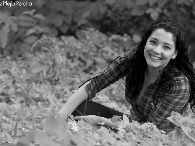 In this picture there is a woman sitting and smiling. At the back there are plants. At the bottom...