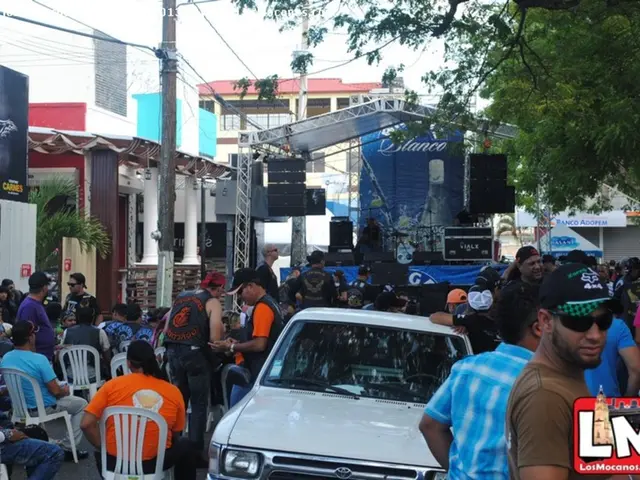 In this image there are few people sitting on the chairs and few are standing. In the center there...