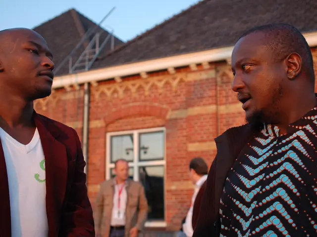 In this picture there are two African boys standing and discussing something. Behind there is a red...